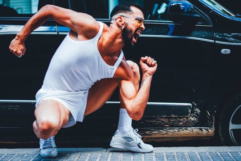 Víctor Óscar Juaranz - En posición de cuclillas con traje corto de color blanco y coche negro de fondo - Horizontal - Color