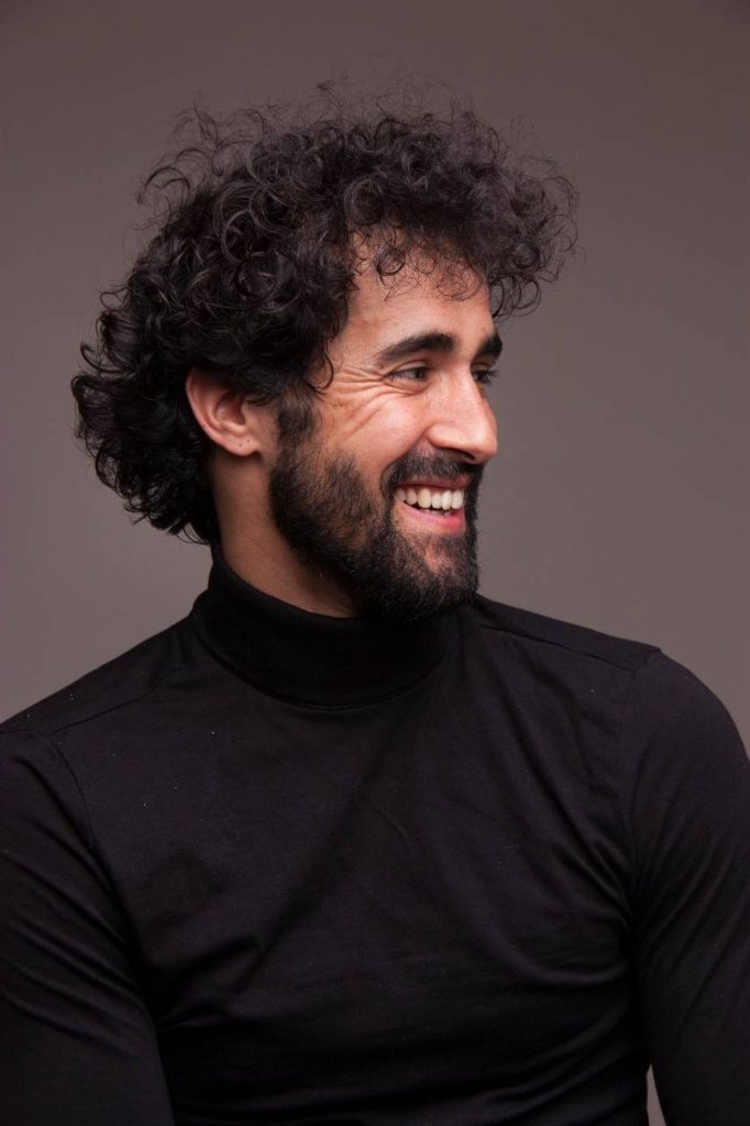 Víctor Óscar Juaranz - Plano medio de perfil con gesto sonriente y jersey negro de cuello cisne - Vertical - Color